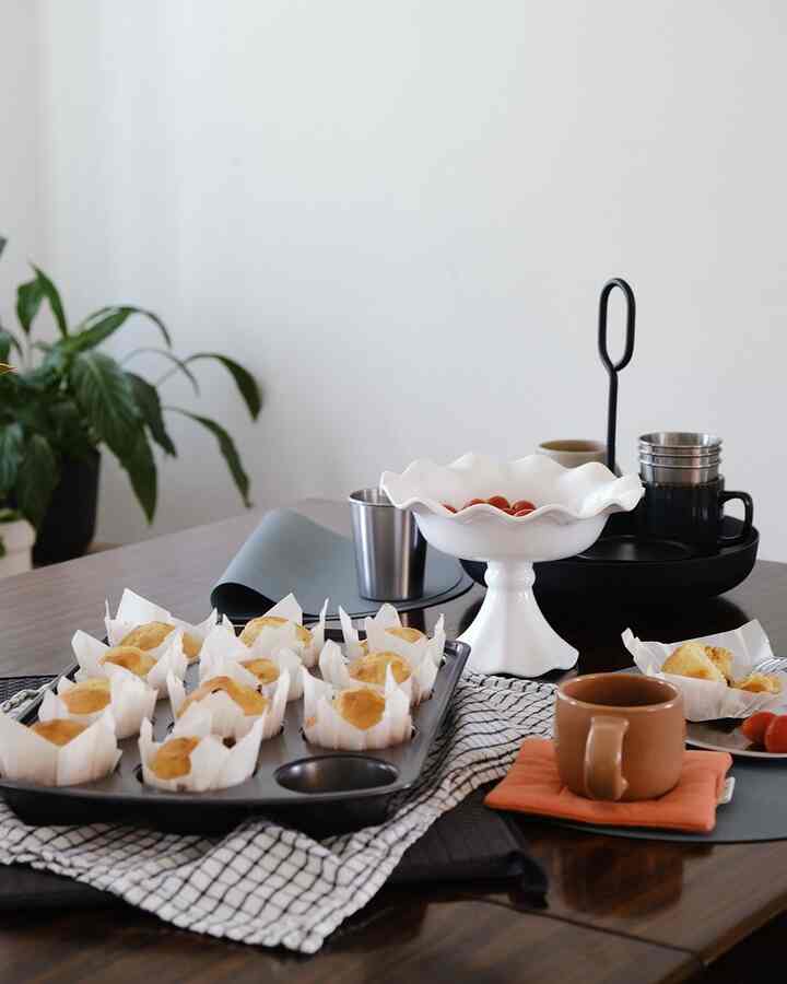 Warm table setting with home cafe mugs, muffins, and silver cups on brown wood table against a white wall