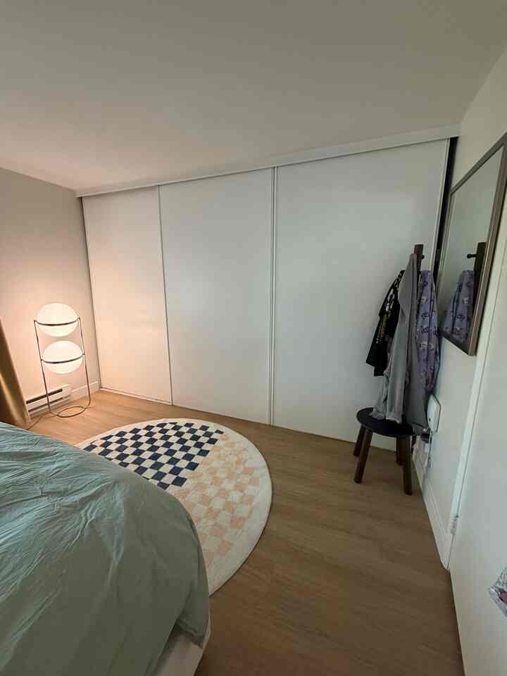 Bedroom with beige wood flooring and white sliding doors, featuring a circular checkered rug and a glowing floor lamp creating a cozy atmosphere