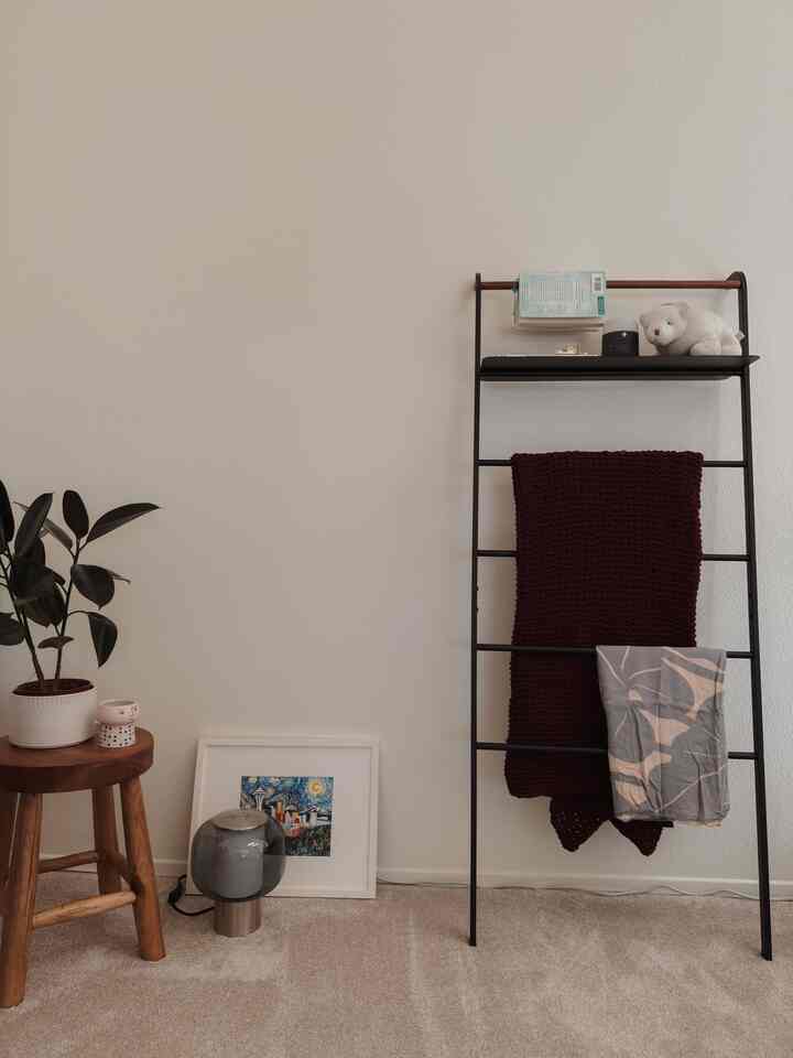 Beige-toned living room corner featuring a black metal blanket ladder, wooden stool, and a potted plant with a calm atmosphere