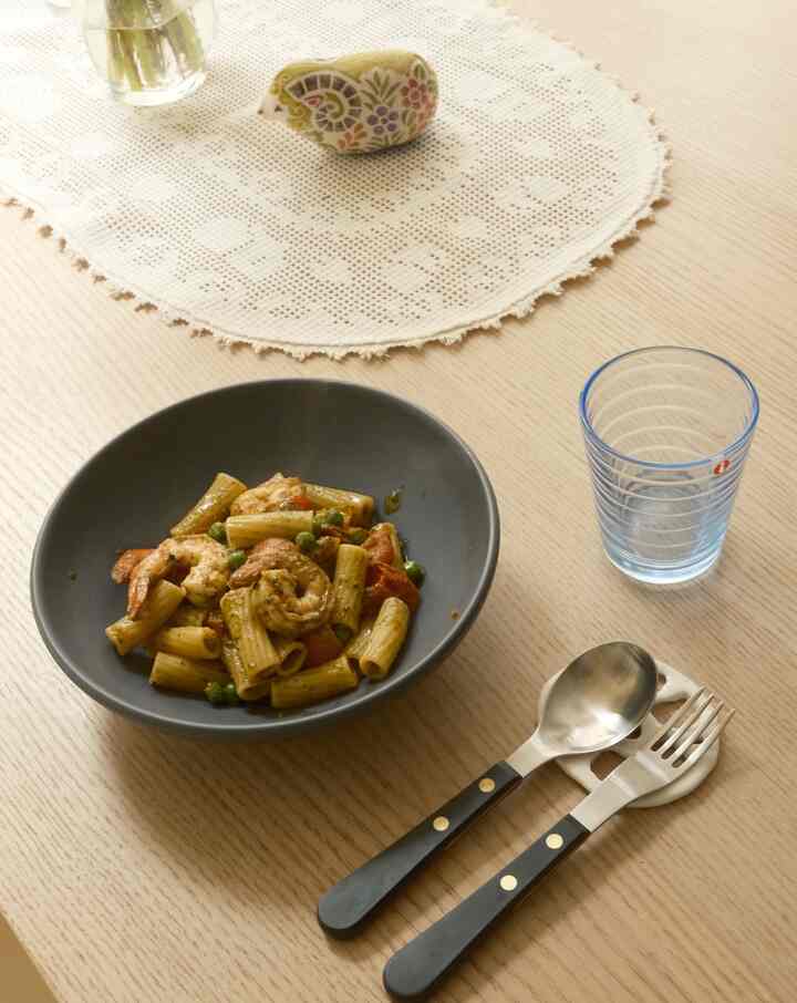 Natural wood-tone dining table featuring a plate of pasta, spoon and fork, and a blue-tinted glass in a simple dining setting