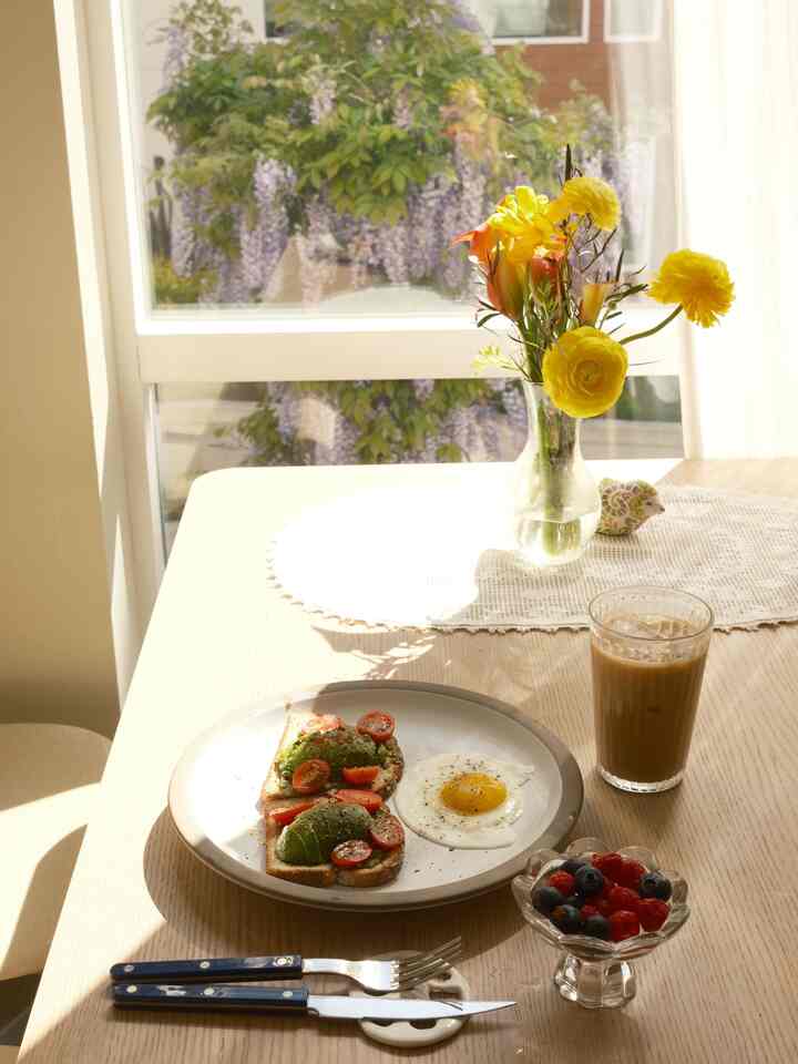 Cozy home cafe setting on a bright wood tone dining table featuring white lace table runner, yellow flower vase, avocado toast, fried egg, and iced coffee