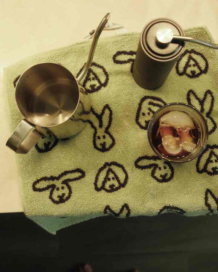 Green towel with black bunny patterns on countertop featuring a stainless steel coffee kettle, hand grinder, and iced coffee cup