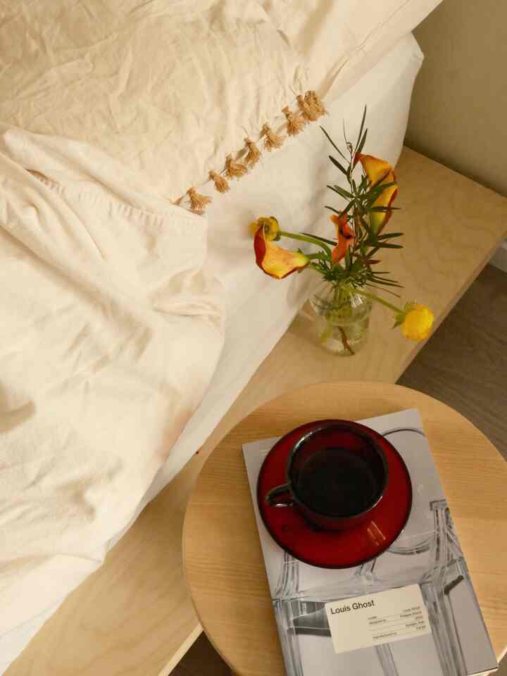Wood tone bedroom with white bedding, featuring a stool holding a coffee cup and vase creating a cozy atmosphere